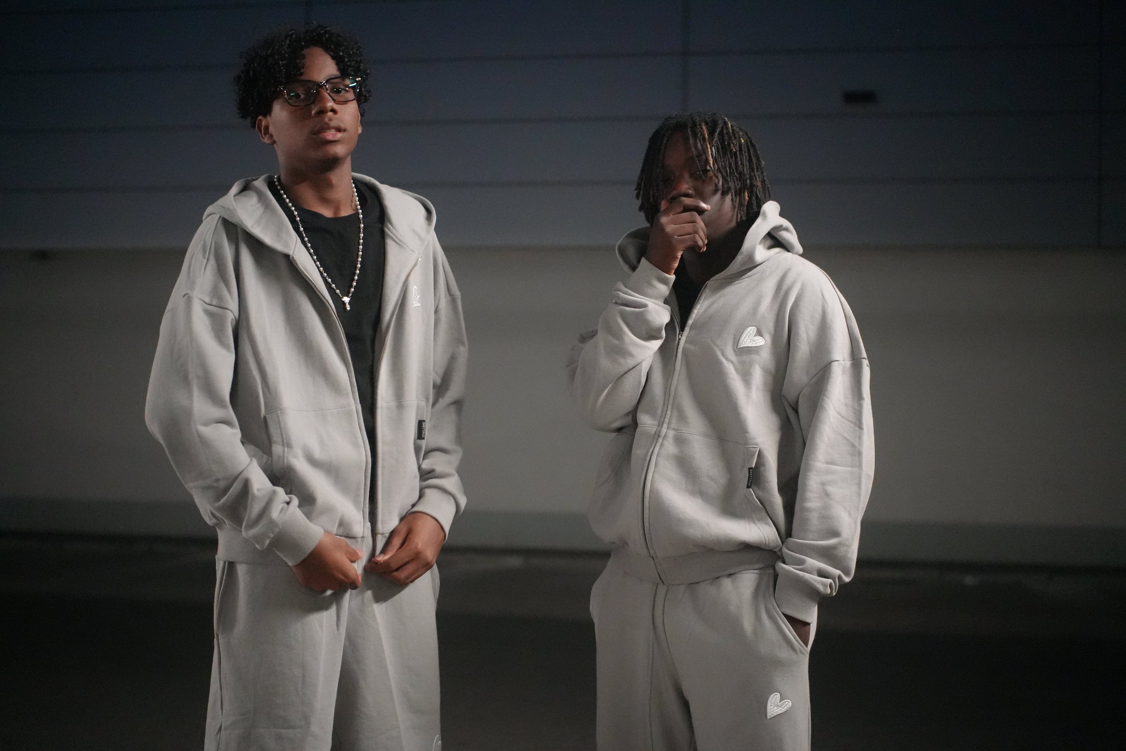 Two individuals wearing gray Infinity tracksuits by Baettage in a dark indoor setting
