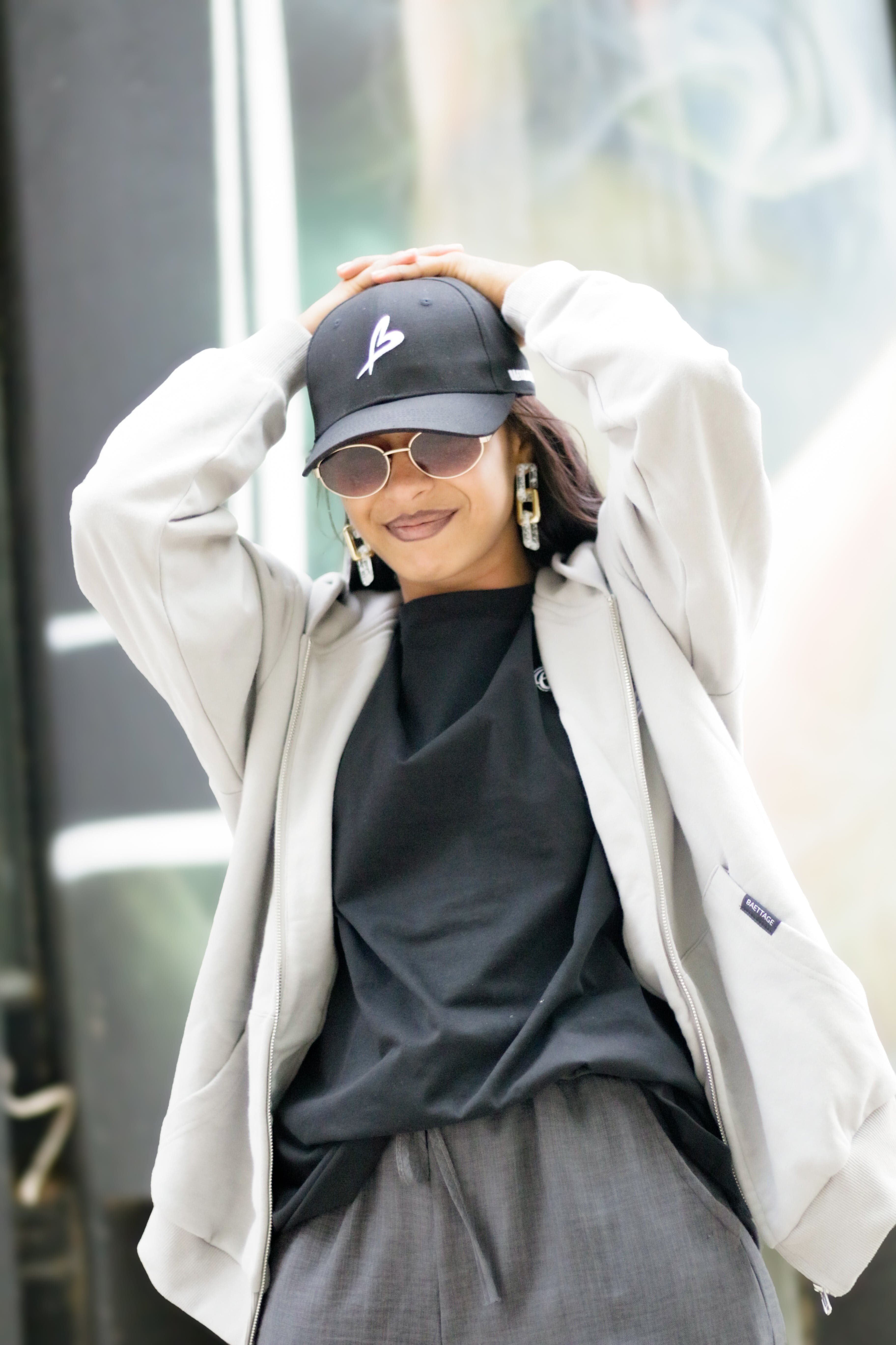 Person wearing a cap, sunglasses, and a light-colored coat outdoors.